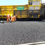 Robotnicy w pomarańczowych strojach układają świeży asfalt przed dużą żółtą układarką drogową