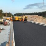 Robotnicy układają świeży asfalt na moście przy użyciu rozściełacza, na tle lasu i placu budowy