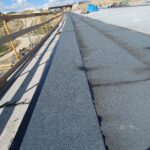 Betonowy most w budowie z ciemną izolacją, barierkami po lewej i nasypami pod błękitnym niebem