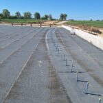 Płaska betonowa płyta budynku w budowie z rzędami kotew, na tle pól, drogi gruntowej i bezchmurnego nieba