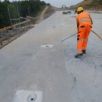 Pracownik w pomarańczowym stroju czyści betonową nawierzchnię budowanego mostu, w tle koparka i skarpy ziemne