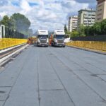 Prace drogowe na moście: dwa ciężarówki i robotnicy układają nawierzchnię między żółtymi barierkami