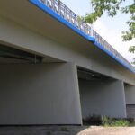 Betonowy most z niebieską krawędzią nad wąskim kanałem, widziany od spodu, z gałęziami drzewa po prawej