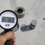 Ręczny miernik siły pokazujący 8,0 kN podczas testu na jasnej betonowej powierzchni z metalowymi elementami