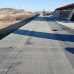 Plac budowy wiaduktu z betonową nawierzchnią, składowanymi zbrojeniami i pagórkowatym krajobrazem w tle