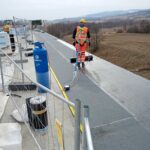 Robotnik w kamizelce i kasku wykonuje prace hydroizolacyjne na dachu, obok niebieskiej butli z gazem