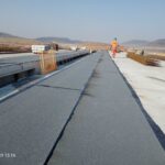 Pracownik w kasku idzie po betonowej konstrukcji na placu budowy, z prętami zbrojeniowymi i wzgórzami w tle
