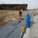 Pracownik budowlany w kasku zgrzewa papę na dachu lub pomoście, obok stoi niebieska butla gazowa