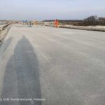 Robotnicy w pomarańczowych strojach pracują na szerokiej betonowej jezdni w budowie, w tle pola i drzewa