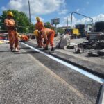 Robotnicy w kaskach montują metalową dylatację na remontowanej jezdni, obok ogrodzenia i samochodów serwisowych