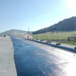 Pracownik nakłada niebieską powłokę na szerokiej betonowej nawierzchni, z polami i zalesionym wzgórzem w tle