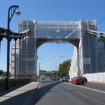 Most nad jezdnią owinięty rusztowaniem i białą folią podczas remontu, pod nim jadą samochody