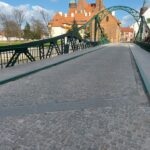 Zielony stalowy most nad rzeką prowadzący do zabytkowych budynków i kościoła pod częściowo zachmurzonym niebem