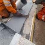 Pracownicy w odzieży ochronnej wylewają i wyrównują mokry beton w długiej formie