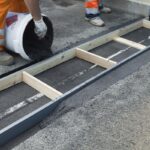 Pracownik w pomarańczowym stroju wylewa masę asfaltową do drewnianej formy na krawędzi chodnika
