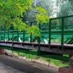 Zielony stalowy most z drewnianym pomostem wśród drzew, oparty na ceglanych filarach
