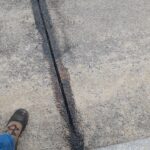 Szczelina dylatacyjna w betonowym chodniku wypełniona czarną masą, widoczne czubki dwóch butów