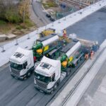 Dwie ciężarówki z maszynami układają asfalt na nowo budowanym moście, obok pracują robotnicy w kamizelkach