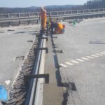 Dwaj robotnicy w pomarańczowych strojach naprawiają szczelinę dylatacyjną na moście