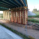 Pod mostem trwa remont podpory; widoczne czerwono-żółte rusztowanie, ogrodzony plac budowy i zielony teren wokół