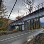 Stalowy most w remoncie nad drogą, z dźwigiem i pracownikami, wśród drzew pod pochmurnym niebem