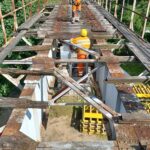Pracownicy w odzieży ochronnej remontują zniszczony drewniano-stalowy most nad zielonym terenem