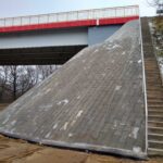 Most z czerwonym bokiem nad stromym umocnionym nasypem, obok wąskie betonowe schody na skarpie