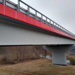 Nowoczesny most z czerwonymi panelami bocznymi nad rzeką, widziany od spodu w pochmurny dzień