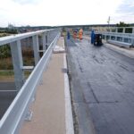 Pracownicy wykonują prace drogowe na nowym wiadukcie z metalowymi barierkami nad okolicznym miasteczkiem