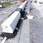 Pracownicy w pomarańczowych strojach montują długi betonowy krawężnik na dachu, obok leżą narzędzia i żwir
