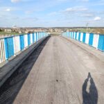 Remontowany most z nieutwardzoną jezdnią i niebiesko-białymi barierkami, z widokiem na wieś pod błękitnym niebem
