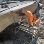 Dwóch robotników na rusztowaniu naprawia betonowy przepust pod drogą w wykopie