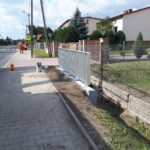 Robotnicy w pomarańczowych strojach pracują przy chodniku na osiedlowej ulicy obok nowo montowanego białego ogrodzenia