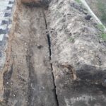 Wąski wykop wzdłuż kostki brukowej, z pękniętą ziemią i odsłoniętym podłożem obok skarpy