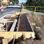 Prace drogowe przy chodniku: zbrojenie w drewnianym szalunku obok jezdni, z barierkami i oznaczeniem objazdu