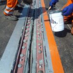 Dwaj robotnicy w pomarańczowych strojach uszczelniają metalową szczelinę na nawierzchni za pomocą narzędzi i masy