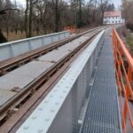 Most kolejowy z szarych stalowych belek i pomarańczową barierką nad strumieniem, prowadzący do białego budynku