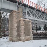 Stalowy most na kamiennym filarze nad strumieniem w zimowym krajobrazie, z pomarańczowymi barierkami