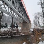 Widok od spodu na stalowy most nad strumieniem w zimowym krajobrazie, z kamiennymi filarami i nagimi drzewami