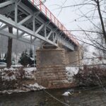 Stalowy most z pomarańczowymi barierkami nad strumieniem w zimowym, lekko ośnieżonym lesie