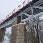Stalowy most kratownicowy na kamiennych filarach zimą, widziany od spodu na tle nagich drzew