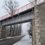 Stalowy most z czerwonymi barierkami nad drogą w zimowym krajobrazie, lekki opad śniegu