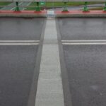 Mokra jezdnia na moście z jasnym pasem pośrodku, zieloną barierką i skrzyżowaniem widocznym w tle