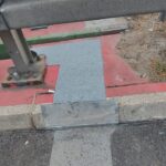 Bariera drogowa przy krawężniku z prowizoryczną metalową nakładką na pękniętej czerwonej nawierzchni