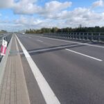Pusta dwupasmowa autostrada na moście, z barierkami po obu stronach i lasem w tle pod częściowo zachmurzonym niebem