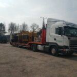Biała ciężarówka Scania Tarcopol z naczepą niskopodwoziową przewożąca dużą żółtą maszynę na placu