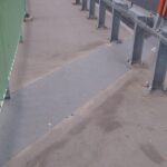 Wąski chodnik na moście z metalowymi barierkami i szarą antypoślizgową rampą przy jezdni